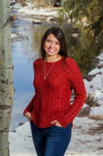 Ana has long, dark hair and is wearing a red sweater and jeans as she stands in front of a snowy lake in winter.