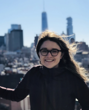 Kendall has long, dark windblown hair and circular glasses. She smiles while standing in front of a city skyline.
