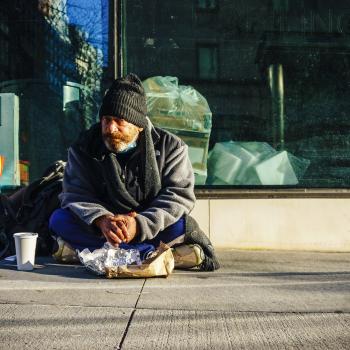Man sitting on a sidewalk with bags and food, appearing unhoused