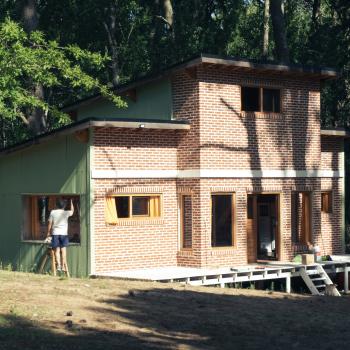 Person standing building a small brick-and-wood house surrounded by trees