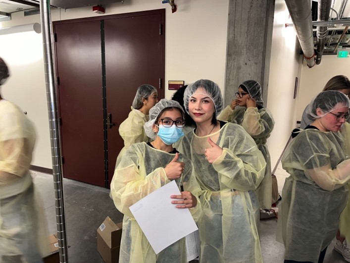 Two people wearing PPE posing and giving thumbs up with additional people in the background